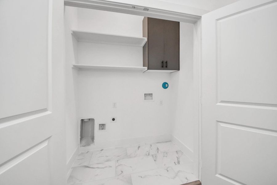 This is a compact laundry room with white walls and a marble like tile floor. It features open shelving and a cabinet for storage, with hookups for a washer and dryer. This is a compact laundry room with white walls and a marble like tile floor. It features open shelving and a cabinet for storage, with hookups for a washer and dryer.