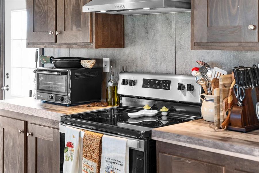 Kitchen with stainless steel electric range, butcher block countertops, dark brown cabinets, and ventilation hood Kitchen with stainless steel electric range, butcher block countertops, dark brown cabinets, and ventilation hood