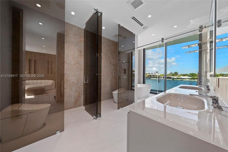 Dual vanity with integrated sinks, full-height sliding glass panels, freestanding tub, recessed lighting, and expansive water frontage