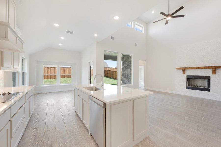 Open kitchen with white cabinets, center island with sink, vaulted ceiling, and brick fireplace in adjacent living area.