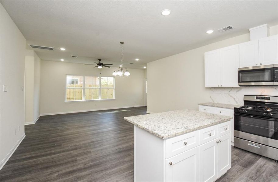 Modern kitchen with white cabinets, granite countertops, stainless steel appliances, and dark wood flooring, leading to a living area with ceiling fans.