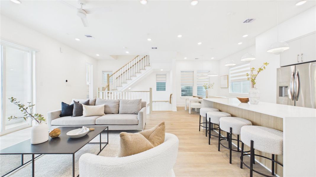 This modern, open-concept living area features a bright and airy design with white walls, light wood flooring, and ample natural light. The spacious layout includes a cozy seating area, a sleek kitchen with a large island and bar stools, and contemporary lighting fixtures. Perfect for entertaining and family gatherings.