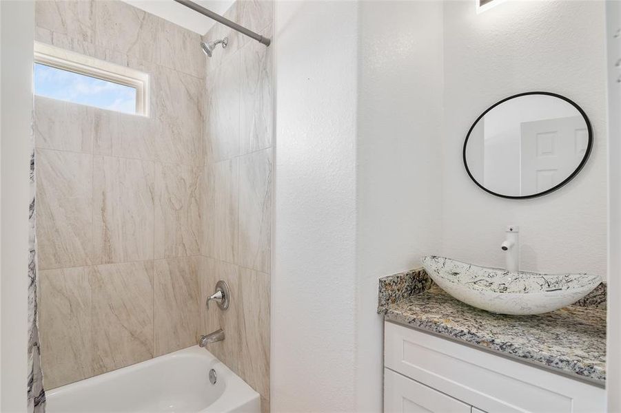 The photo shows a modern secondary  bathroom with a tiled bathtub, a small window for natural light, a unique vessel sink atop a granite countertop, and a circular mirror.