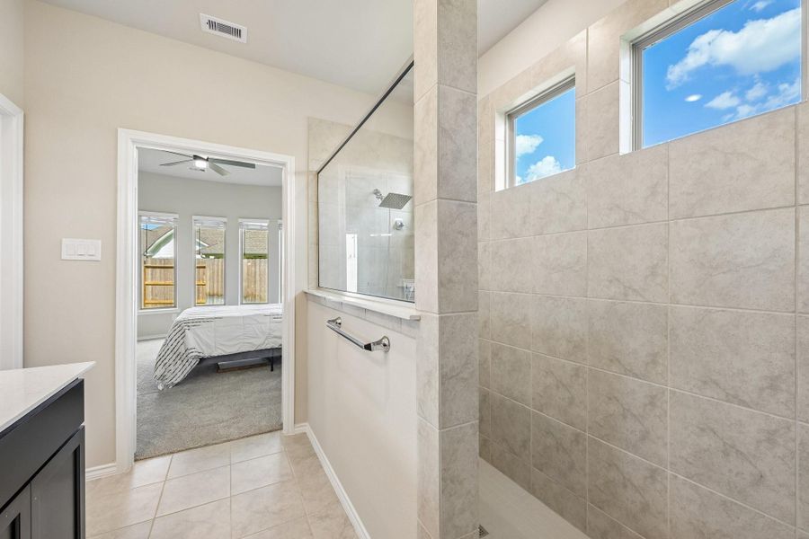 Elegant bathroom with a spacious walk-in shower, featuring sleek tilework and natural light from high windows, seamlessly connected to a cozy bedroom with a ceiling fan and large windows.