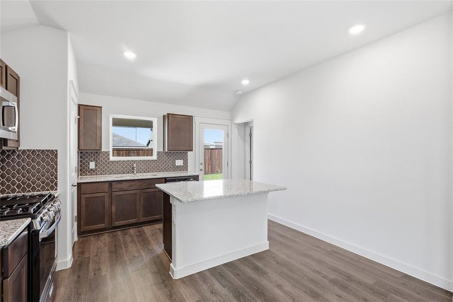 Kitchen with vaulted ceiling, light stone counters, backsplash, gas range oven, and a center island