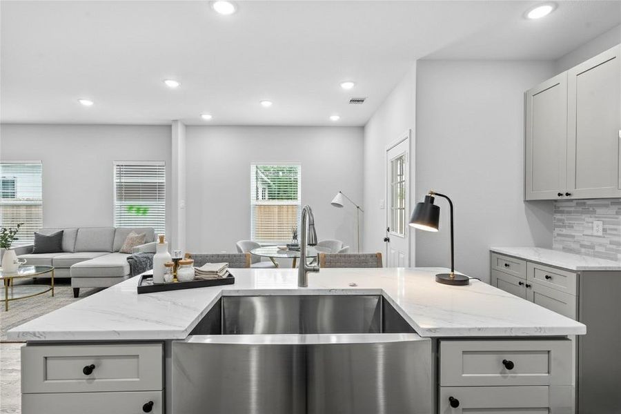 Modern open-concept kitchen and living area with a spacious island, stainless steel sink, and sleek cabinetry. The room features ample natural light from large windows, recessed lighting, and a cozy seating area. **Photo has been staged.**