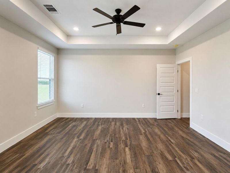 A spacious bedroom features rich wood flooring, a modern ceiling fan, and recessed lighting in a tray ceiling.