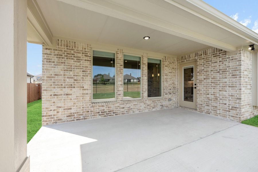 Covered back patio with concrete floor, brick exterior wall, large windows, and glass door overlooking a grassy backyard.