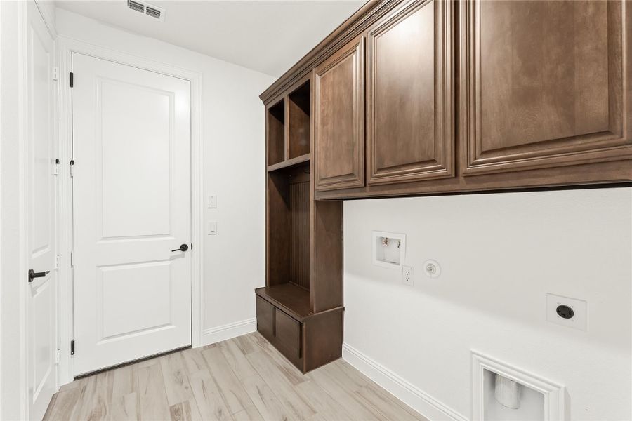 Laundry room with dark wood upper cabinets, open cubbies, and washer and dryer hookups on light hardwood floors Laundry room with dark wood upper cabinets, open cubbies, and washer and dryer hookups on light hardwood floors