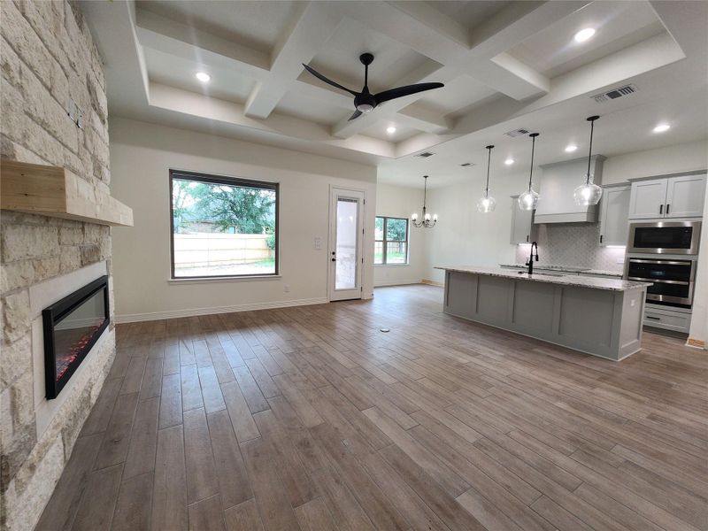 Open-concept living area featuring wood-finish flooring, a stone-clad fireplace with wood mantel, a coffered ceiling with recessed lighting and ceiling fan, a large window, and a kitchen island with pendant lighting