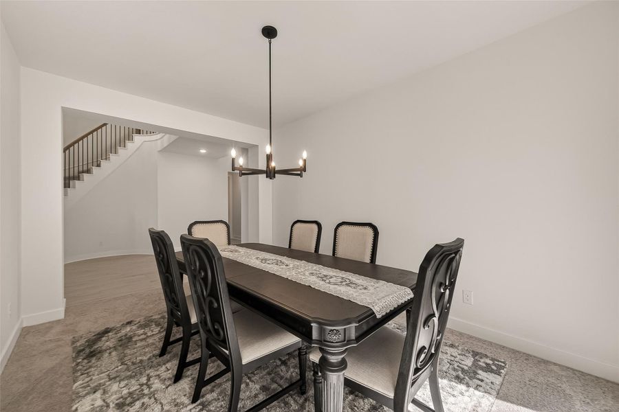 A modern dining room with neutral tones, featuring a dark wood table and six upholstered chairs. A contemporary chandelier hangs above, and the room opens to a staircase with a sleek railing.