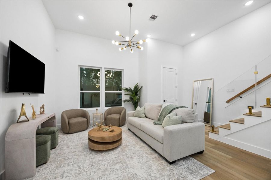 This modern living room features a cozy beige sofa, stylish armchairs, and a round wooden coffee table on a textured rug. A sleek TV is mounted on the wall opposite the sofa. The space is brightened by a contemporary chandelier, with stairs featuring glass railings leading up. Large windows and decor accents add elegance and warmth. This modern living room features a cozy beige sofa, stylish armchairs, and a round wooden coffee table on a textured rug. A sleek TV is mounted on the wall opposite the sofa. The space is brightened by a contemporary chandelier, with stairs featuring glass railings leading up. Large windows and decor accents add elegance and warmth.