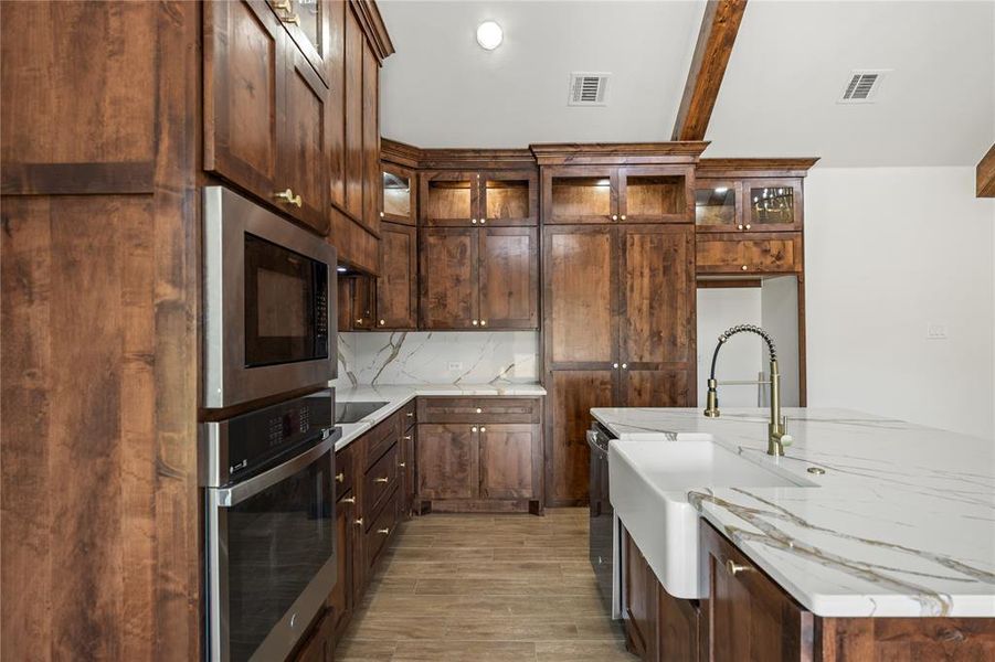 Kitchen with stainless steel appliances, light stone countertops, glass fronted cabinets, light wood-type flooring, and beamed ceiling