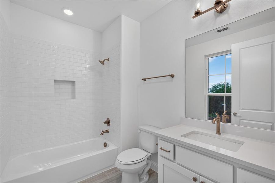 Bathroom featuring a white subway tile shower surround with a built-in niche, a white vanity with an integrated sink and quartz countertop, brushed bronze fixtures, and wood-finish flooring Bathroom featuring a white subway tile shower surround with a built-in niche, a white vanity with an integrated sink and quartz countertop, brushed bronze fixtures, and wood-finish flooring