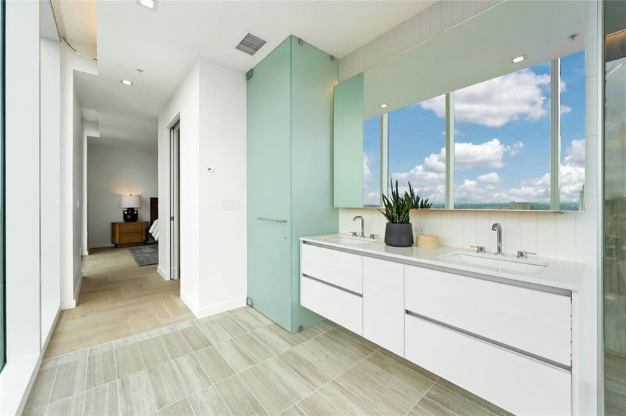 Primary bathroom with a floating double vanity, expansive mirror, and a private water closet tucked behind a frosted glass door. Floor-to-ceiling windows fill the space with natural light and frame skyline views.