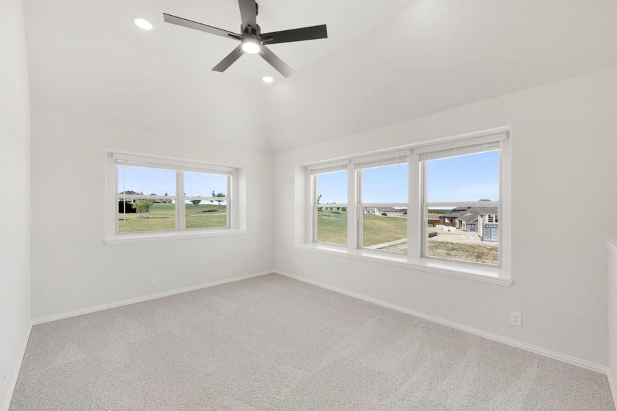 Empty bedroom with vaulted ceiling, ceiling fan, carpet flooring, and large windows overlooking a neighborhood