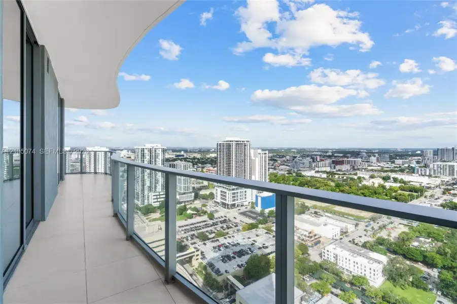 Spacious private balcony featuring large-format tile flooring, a curvilinear overhead architectural element, and a full-height glass railing