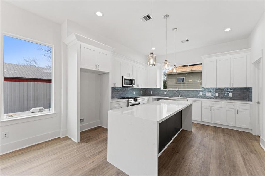 This modern kitchen features an open layout with white cabinetry, a sleek island with a quartz countertop, stainless steel appliances, and stylish pendant lighting. The space is bright and inviting, with wood-look flooring and a large window for natural light.