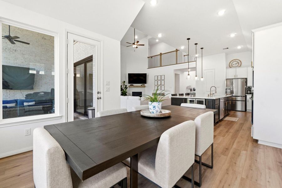 This photo showcases a modern, open-concept kitchen and dining area with high ceilings, stylish light fixtures, and a cozy living space. The room is bright and airy, featuring  a spacious kitchen island, and an adjacent outdoor view through large windows and a glass door.