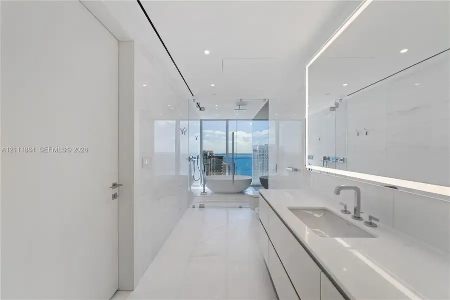 Modern bathroom featuring floor-to-ceiling tiled walls, a floating vanity with a seamless countertop, a large illuminated vanity mirror, a glass-enclosed shower, and a freestanding soaking tub Modern bathroom featuring floor-to-ceiling tiled walls, a floating vanity with a seamless countertop, a large illuminated vanity mirror, a glass-enclosed shower, and a freestanding soaking tub
