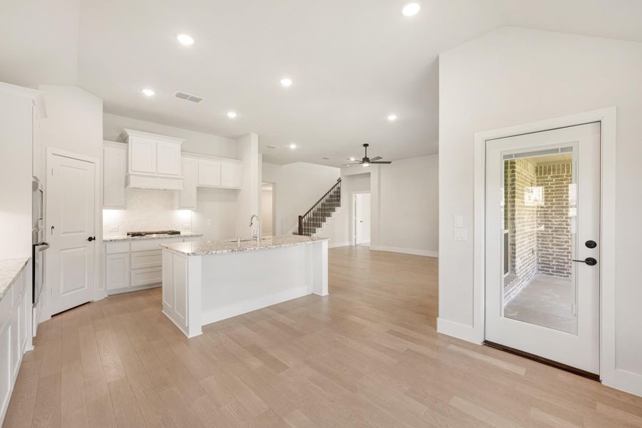 Open-concept kitchen with white cabinets, granite island, and light hardwood floors leading to living area and staircase.