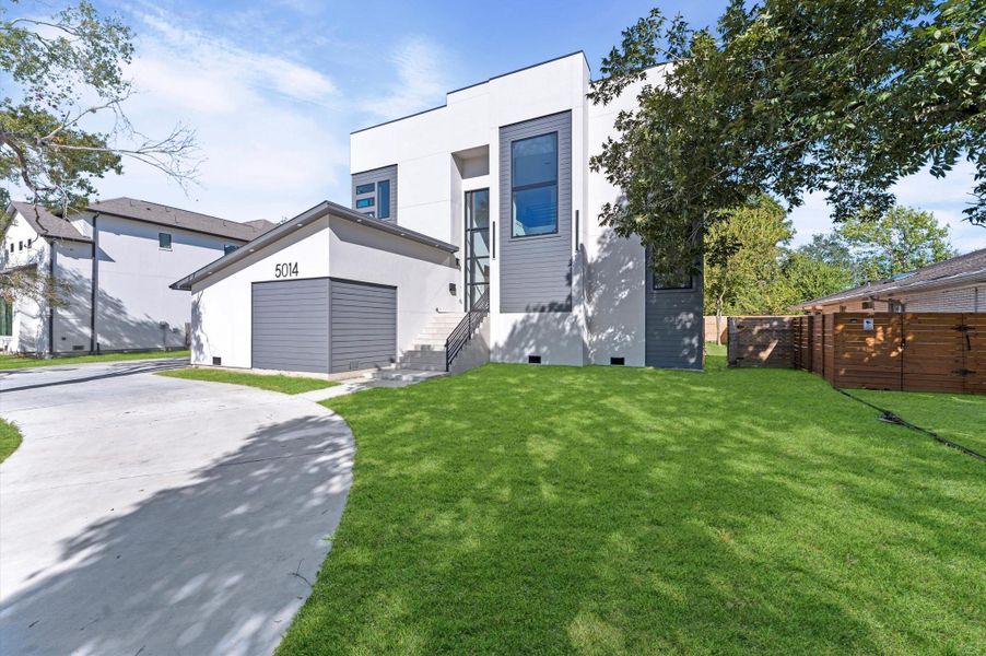 This modern home features a sleek, minimalist design with large windows and a clean white exterior. It has a spacious front yard with lush green grass and a private driveway leading to a two-car garage. The property is surrounded by mature trees, offering a blend of privacy and natural beauty.
