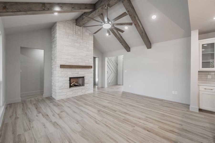 Unfurnished living room featuring a ceiling fan, light wood-type flooring, high vaulted ceiling, a stone fireplace, and beam ceiling
