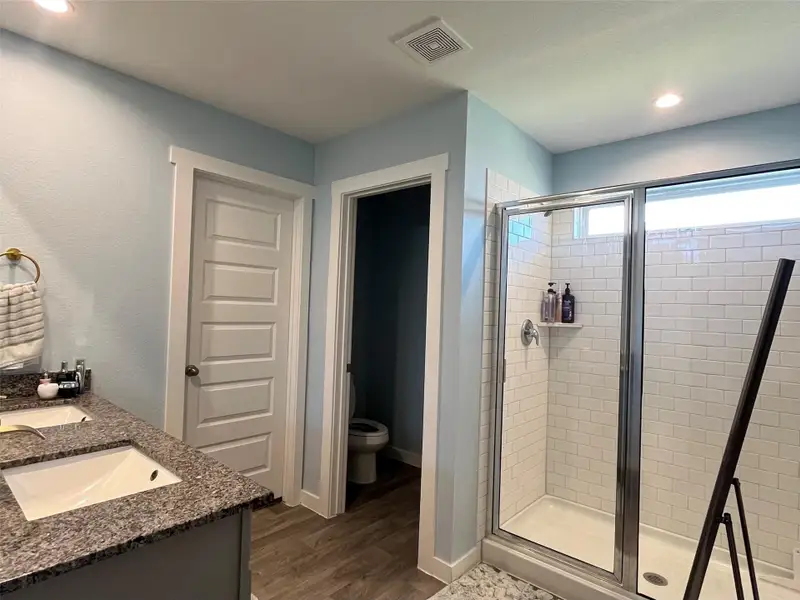 This bathroom features a modern design with a double-sink vanity and granite countertop. It includes a spacious glass-enclosed shower with white subway tiles, a separate toilet area, and wood-look flooring. The soft blue walls and recessed lighting create a bright, inviting atmosphere.