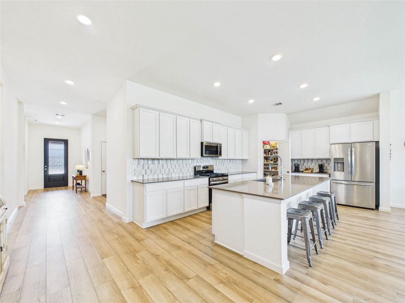 This bright white kitchen is both stylish and functional, featuring abundant cabinetry, quartz countertops, a beautiful tile backsplash, stainless steel appliances, and a corner walk-in pantry. The spacious island with breakfast bar seating offers the perfect spot for casual meals, meal prep, and gathering with family and friends.