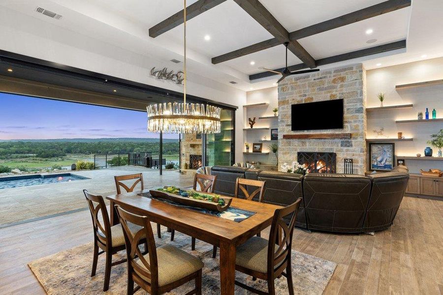 Open-concept living space featuring a retractable wall of glass, a stone-clad fireplace with a hearth, a coffered ceiling with exposed wood beams, a crystal chandelier, and built-in shelving with integrated lighting