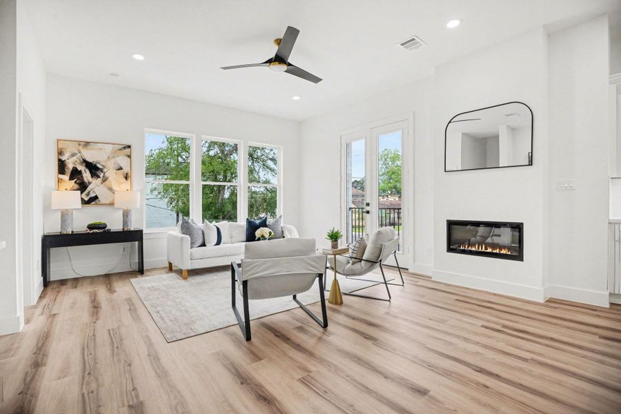 Bright and modern living room with large windows, sleek wood flooring, and a cozy fireplace. It features contemporary furniture, a stylish ceiling fan, and a large wall mirror, creating an inviting and spacious atmosphere.