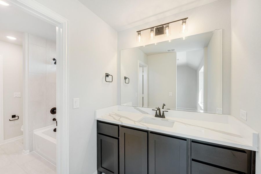 Bathroom with dark vanity cabinet, white quartz countertop, large mirror, and soaking tub visible in background Bathroom with dark vanity cabinet, white quartz countertop, large mirror, and soaking tub visible in background