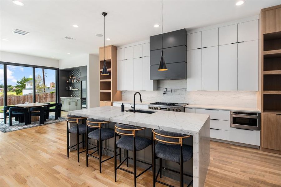 Spacious chef’s kitchen featuring sleek custom cabinetry and a large waterfall island with seating. Premium appliances, designer lighting, and warm wood accents add style and functionality, while the open layout seamlessly connects to the dining area and built-in bar for effortless entertaining.