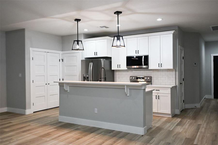 Kitchen featuring appliances with stainless steel finishes, white cabinetry, decorative backsplash, a kitchen island with sink, and hanging light fixtures