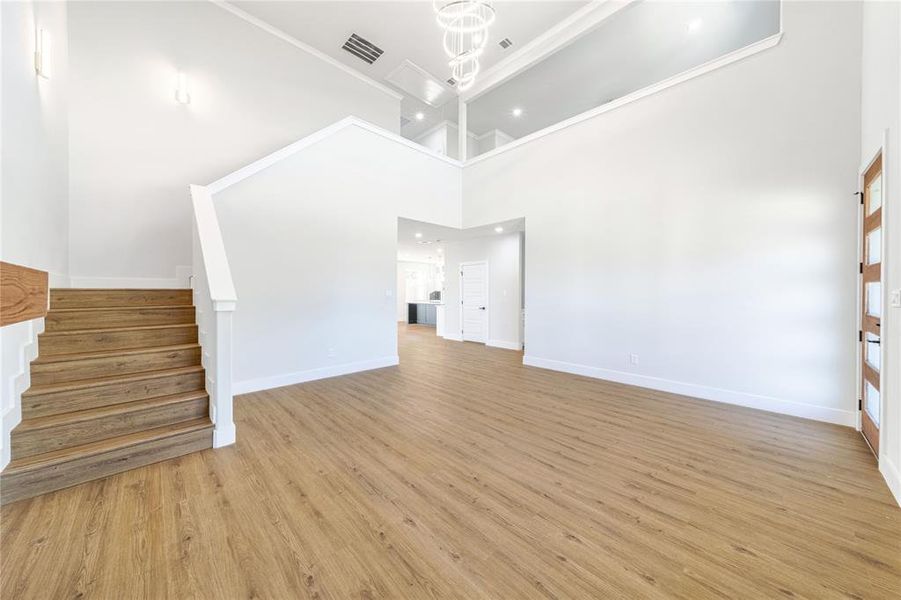 Unfurnished living room with light wood-style floors, a chandelier, a high ceiling, stairs, and ornamental molding Unfurnished living room with light wood-style floors, a chandelier, a high ceiling, stairs, and ornamental molding