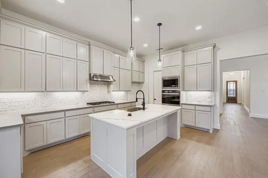 Kitchen with backsplash, light wood-style flooring, a center island with sink, decorative light fixtures, and stainless steel appliances
