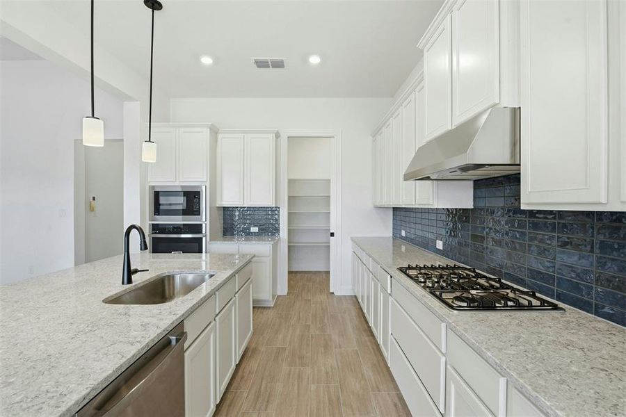 Modern kitchen featuring an island with a stainless steel sink, light-toned countertops, white cabinetry, a gas cooktop with a stainless steel vent hood, and a dark subway tile backsplash