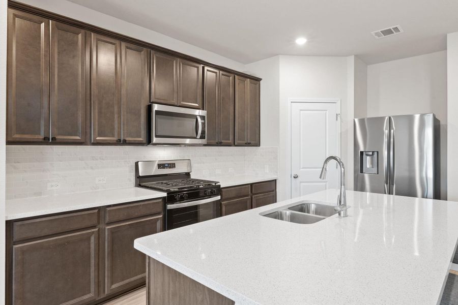 Kitchen with light stone countertops, stainless steel appliances, dark brown cabinets, an island with sink, and decorative backsplash Kitchen with light stone countertops, stainless steel appliances, dark brown cabinets, an island with sink, and decorative backsplash