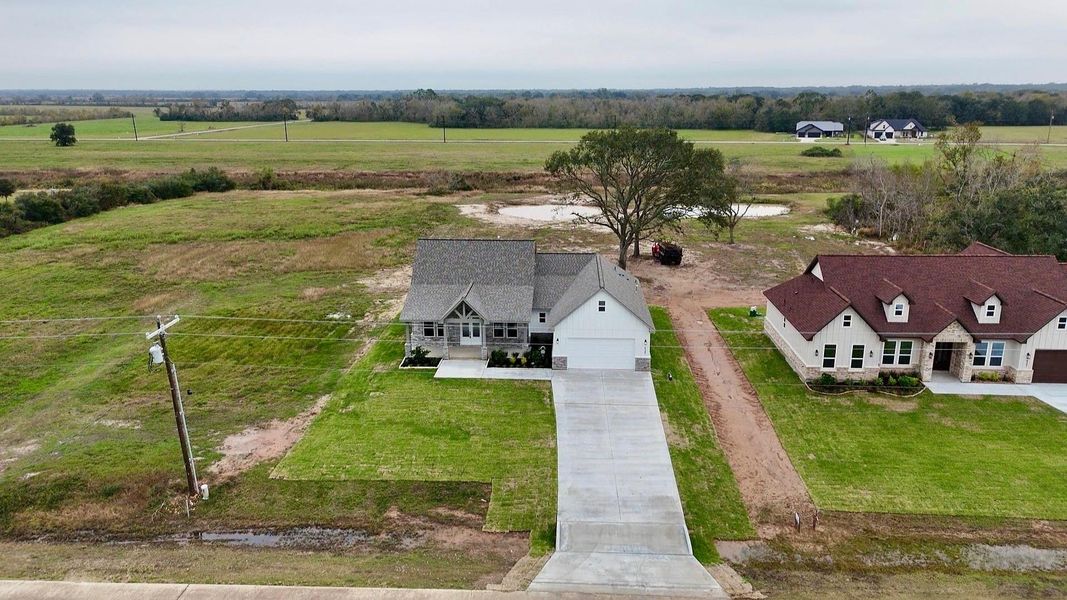 This photo shows a single-family home with a gray roof and a spacious driveway, situated on a large lot. The property has a well-maintained lawn and is adjacent to another similar house. It is surrounded by open fields, offering a sense of privacy and plenty of natural scenery.