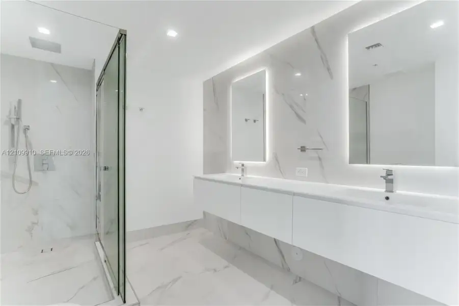 Floating double vanity with dual rectangular sinks, floor-to-ceiling stone-finish tile wall, glass-enclosed walk-in shower with rainfall fixture, and recessed LED-backlit mirrors