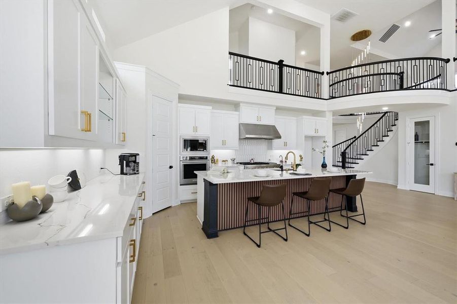 Gourmet kitchen featuring a waterfall island, stainless steel appliances, light wood-finish flooring, white cabinetry with gold-tone hardware, and a sweeping curved staircase