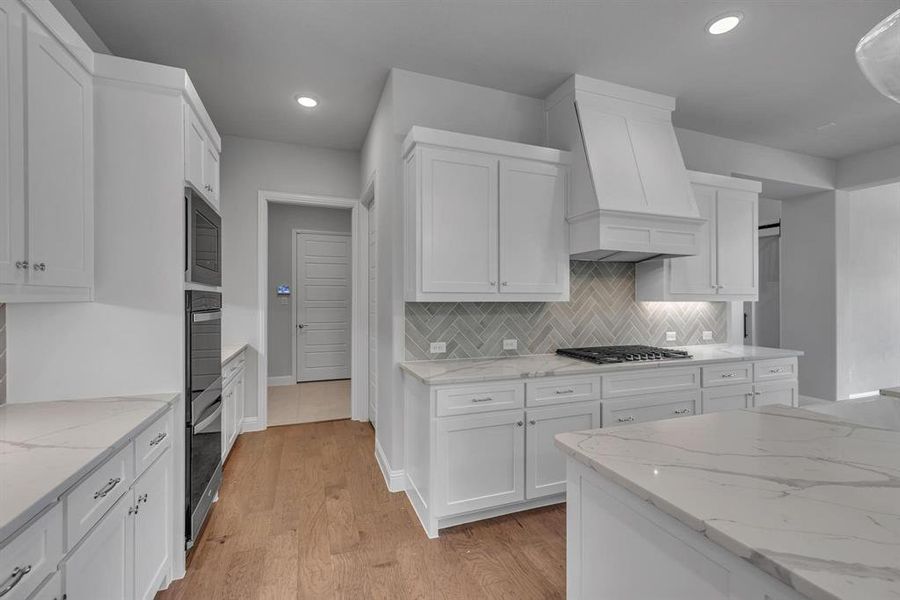 Kitchen with custom range hood, appliances with stainless steel finishes, light stone counters, light wood-type flooring, and white cabinets