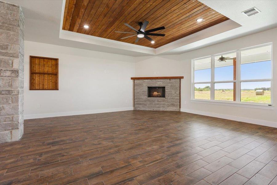 A spacious great room showcases a warm fireplace, a decorative wood-paneled ceiling with a fan, and large windows offering verdant field views.