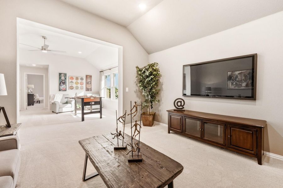 Open living area with vaulted ceiling, wall-mounted TV, wood coffee table, and carpeted floors leading to a game room. Open living area with vaulted ceiling, wall-mounted TV, wood coffee table, and carpeted floors leading to a game room.