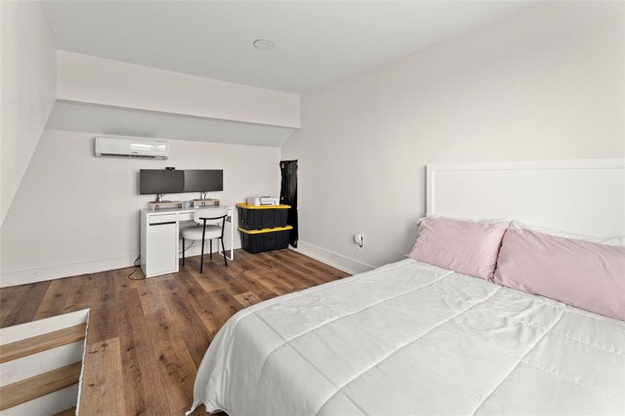 Room featuring wood-finish flooring, a white headboard, a wall-mounted ductless mini-split system, and a recessed ceiling light Room featuring wood-finish flooring, a white headboard, a wall-mounted ductless mini-split system, and a recessed ceiling light