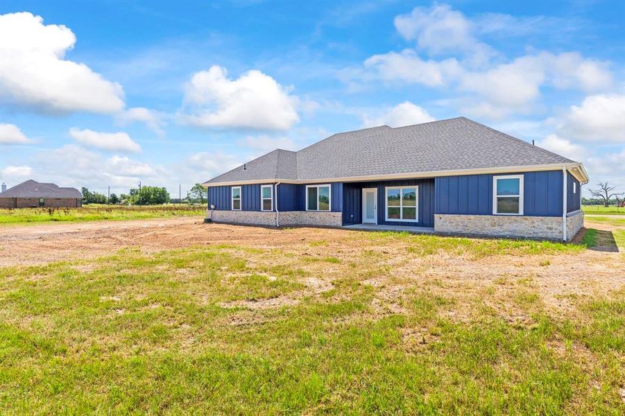 Rear view of house featuring board and batten siding, roof with shingles, stone siding, and a lawn