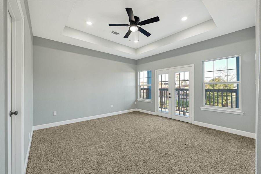 This spacious Primary bedroom features a tray ceiling with recessed lighting and a ceiling fan. It has neutral-toned walls and carpeting, with ample natural light streaming through double French doors and two windows, leading to a balcony. Perfect for a bright, airy living space.