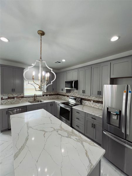 Kitchen featuring a sink, a notable chandelier, decorative backsplash, gray cabinets, and appliances with stainless steel finishes Kitchen featuring a sink, a notable chandelier, decorative backsplash, gray cabinets, and appliances with stainless steel finishes