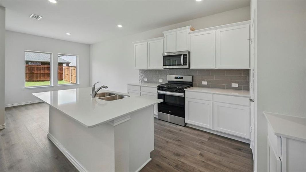 Modern kitchen featuring an expansive center island with an undermount double-basin sink, white shaker-style cabinetry, stainless steel appliances, a subway tile backsplash, and wood-finish flooring Modern kitchen featuring an expansive center island with an undermount double-basin sink, white shaker-style cabinetry, stainless steel appliances, a subway tile backsplash, and wood-finish flooring