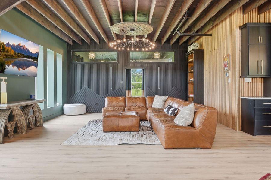 This modern living room features a stylish exposed beam ceiling, a cozy leather sectional, and a unique light fixture. Large windows offer natural light, and a wall-mounted TV enhances entertainment options. The space is accented with wood and black cabinetry for a contemporary feel.